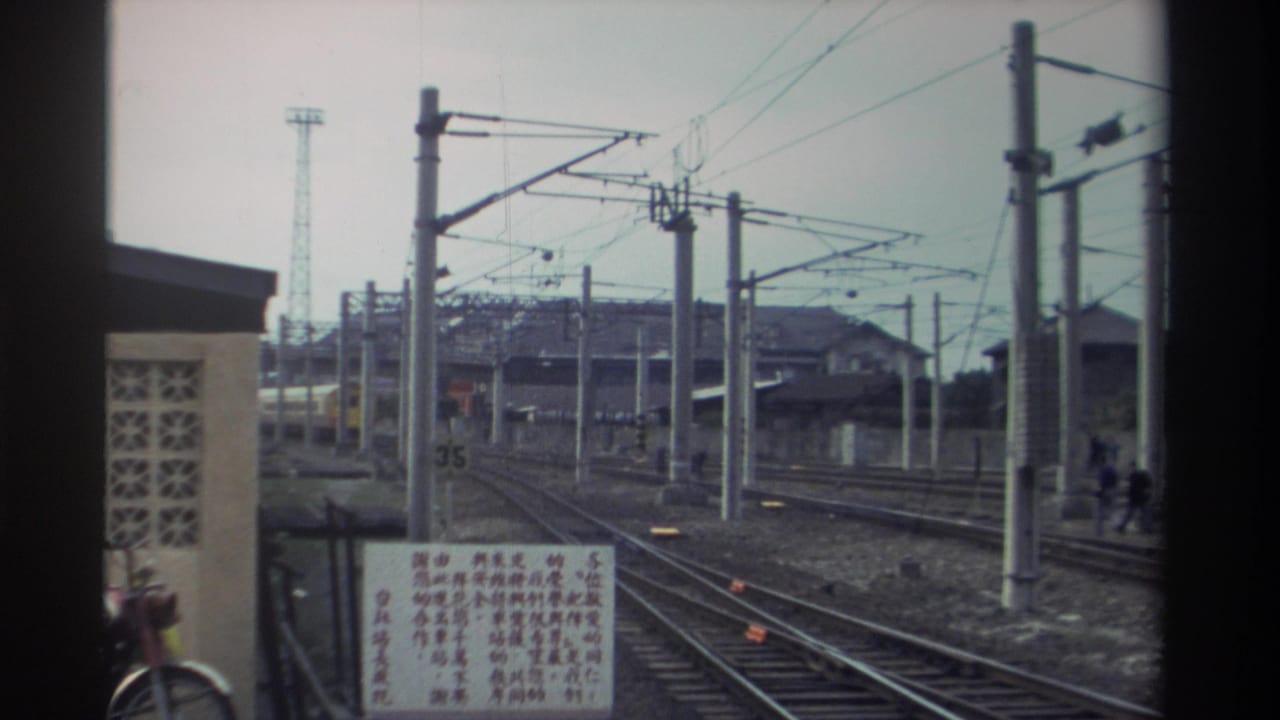 View toward receding railway tracks from a platform or slow-moving train, with a Chinese-character sign at left, concrete catenary poles, and a lattice tower in the distance.
