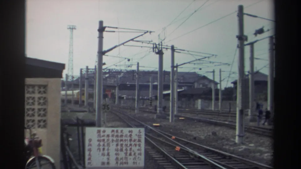1982 Taiwan railway station approach — Chinese-character sign at left, catenary-strung concrete poles, lattice tower, and distant figures on or near the tracks.
