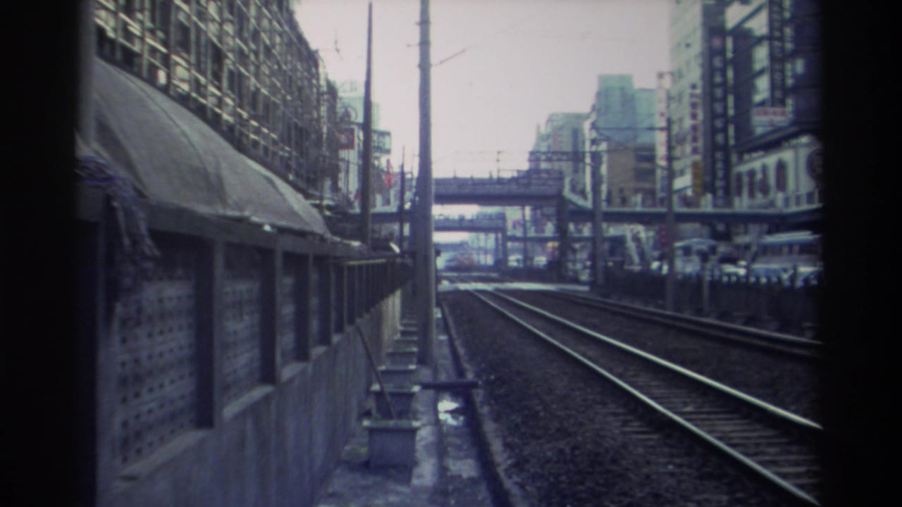 Train-level view along urban tracks with a concrete canopy shelter at left and multi-story commercial buildings and a pedestrian overpass at right.