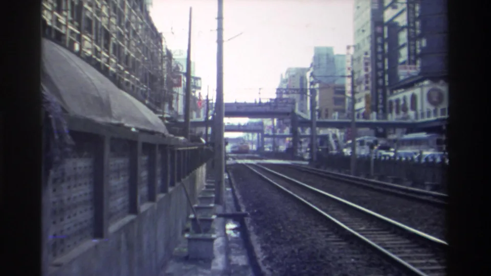 Continuing 1982 Taiwan rail journey — concrete shelter canopy, catenary poles, high-rise buildings with Chinese signage, and a pedestrian overpass visible ahead.