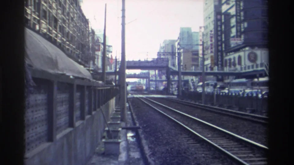 Final frames of 1982 Taiwan rail journey — urban corridor with catenary poles, commercial buildings with Chinese signage, pedestrian overpass, and receding parallel tracks.