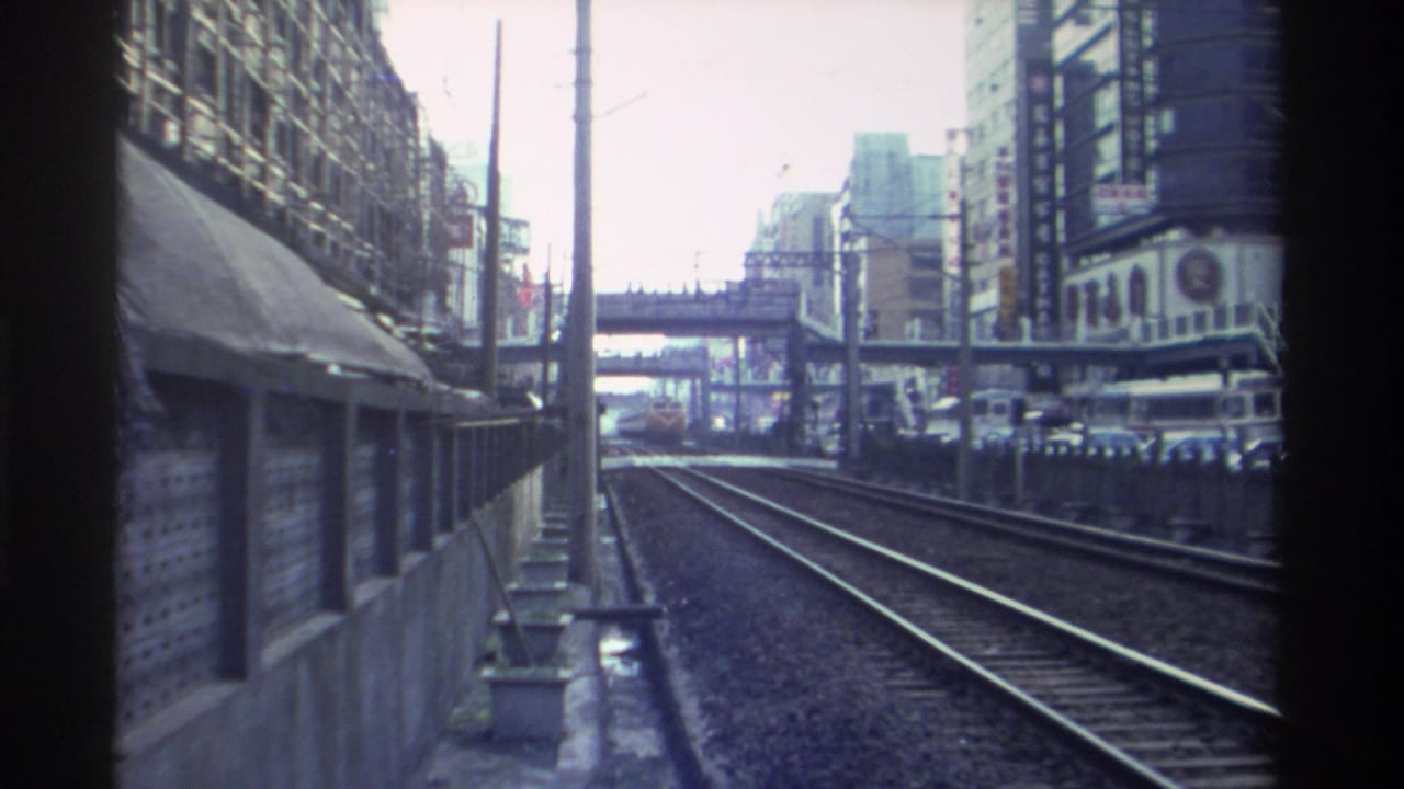 Railway POV from a moving train through a dense urban corridor, Taiwan circa 1982; overhead pedestrian bridge and commercial buildings frame the tracks.