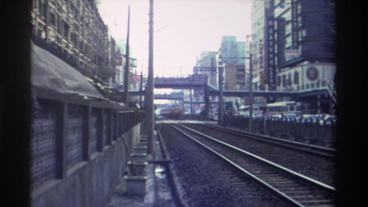 Continuing train-window POV along urban railway, Taiwan circa 1982; pedestrian overpass visible ahead, dense commercial streetscape on both sides.