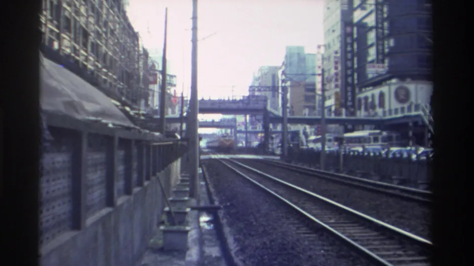 Continuing train-window POV along urban railway, Taiwan circa 1982; pedestrian overpass visible ahead, dense commercial streetscape on both sides.