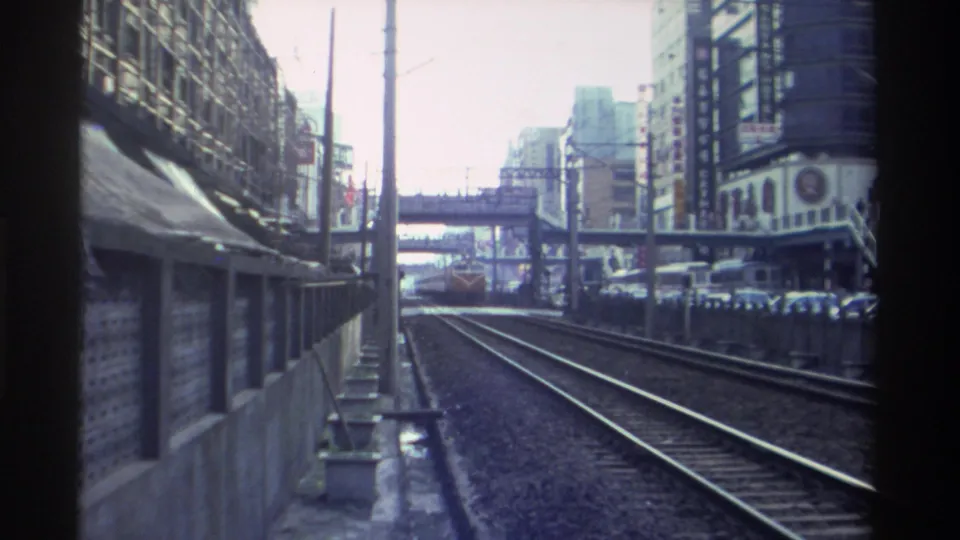 Train-window POV, Taiwan circa 1982; a locomotive approaches on the tracks ahead, pedestrian overpass spanning the corridor, commercial buildings on both sides.