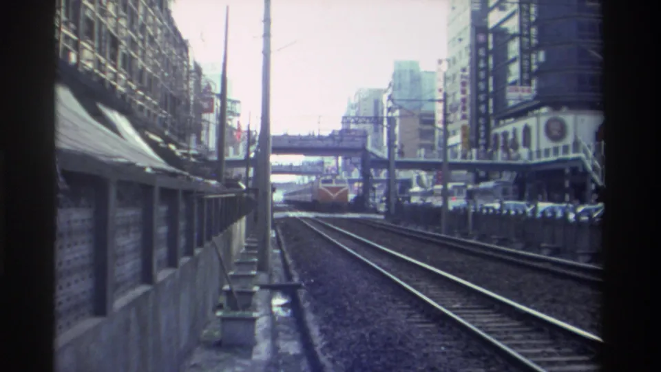Approaching locomotive on urban railway, Taiwan circa 1982; pedestrian overpass and multi-story commercial buildings frame the tracks on both sides.