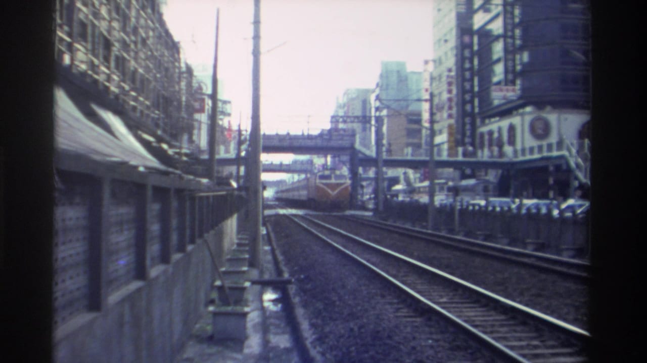 Low-angle 8mm view down a Taiwan railway corridor circa 1982, orange-and-cream diesel locomotive approaching in the distance between concrete barriers and flanking high-rises.