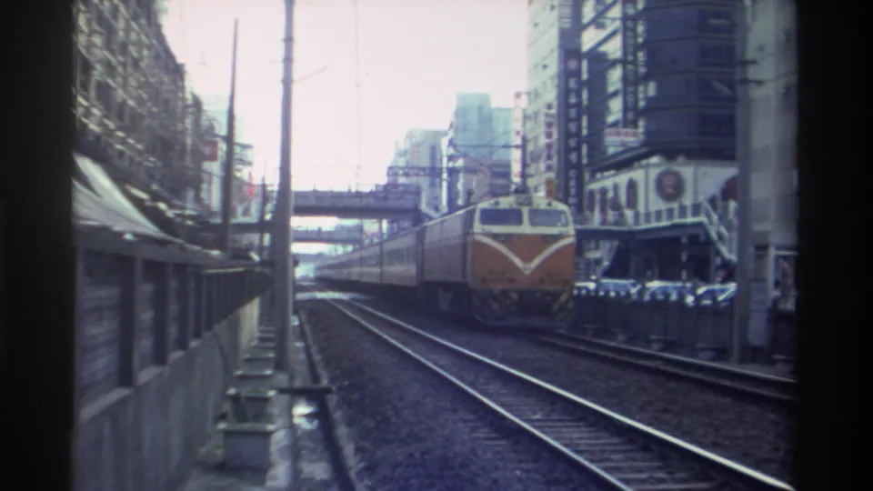 8mm footage from Taiwan circa 1982 — orange diesel locomotive at closer range, urban commercial buildings and a pedestrian footbridge framing the track corridor.
