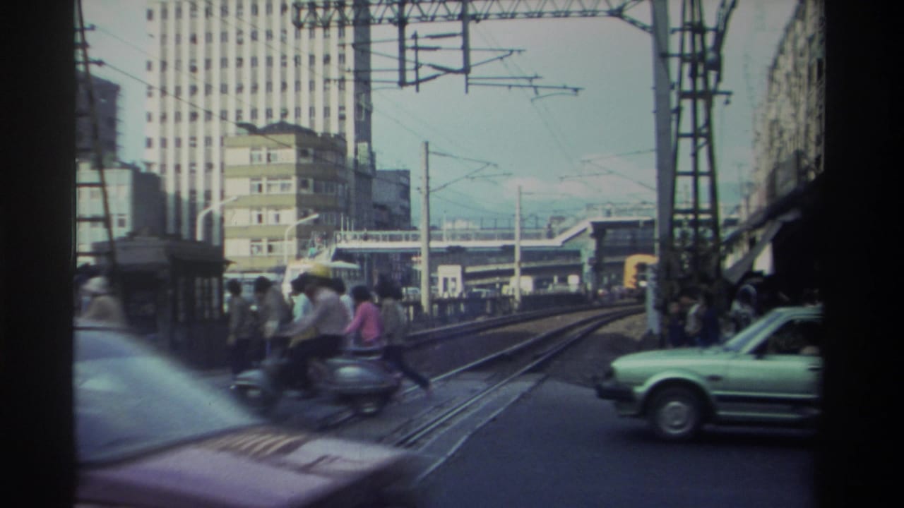 Urban street in Taiwan, 1982 — pedestrians, moped rider, pale-green sedan, tram tracks, and overhead electric wires, shot from a moving vehicle.