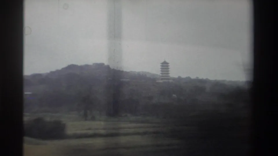 1982 Taiwan, 8mm film — multi-tiered pagoda on tree-covered hillside viewed through a car window; cultivated fields in foreground, overcast sky.
