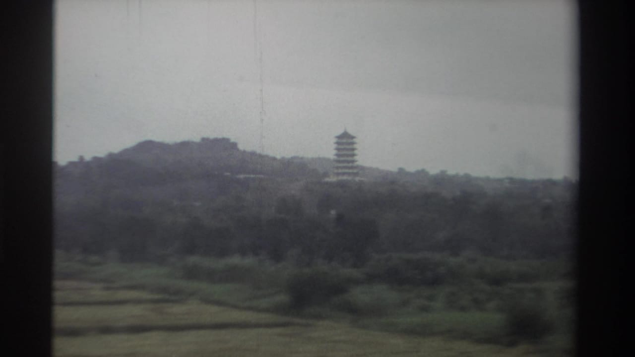1982 Taiwan, 8mm film — pagoda on forested hillside as car continues past; cultivated fields and grey sky visible, no window obstruction.