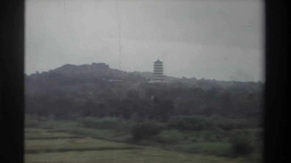 1982 Taiwan, 8mm film — pagoda on forested hillside as car continues past; cultivated fields and grey sky visible, no window obstruction.