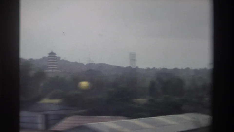 8mm frame from a moving vehicle in Taiwan, circa 1982; pagoda recedes left while a corrugated rooftop, yellow reflection, and a utility tower appear in the foreground.
