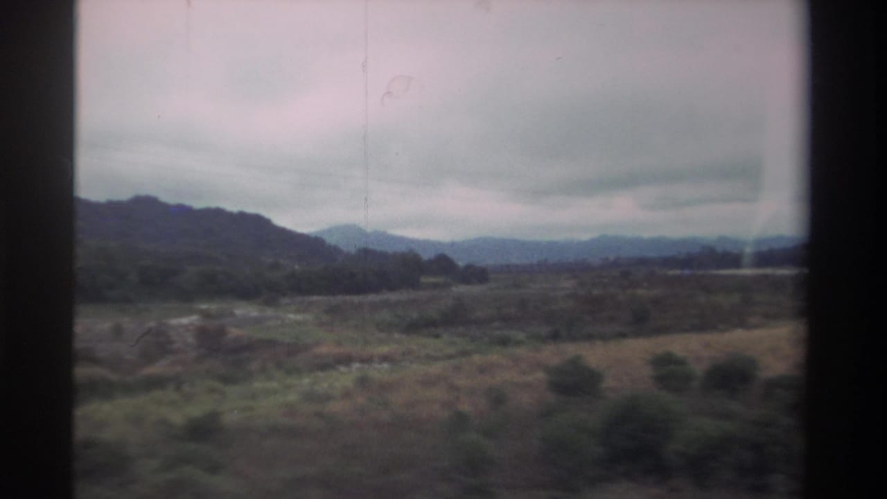 8mm home movie, 1982 Taiwan — dry scrubland and rolling mountains seen through a moving vehicle window under heavy cloud cover.