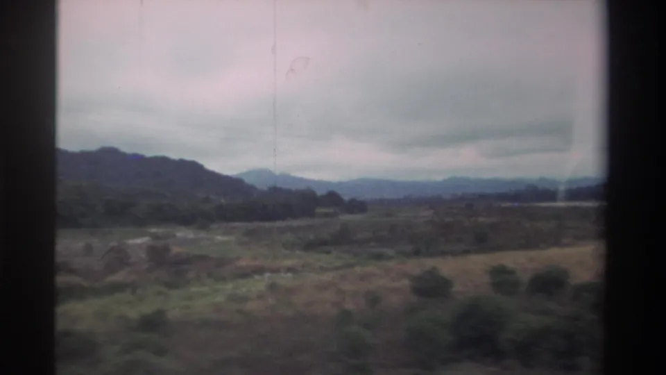 8mm home movie, 1982 Taiwan — dry scrubland and rolling mountains seen through a moving vehicle window under heavy cloud cover.
