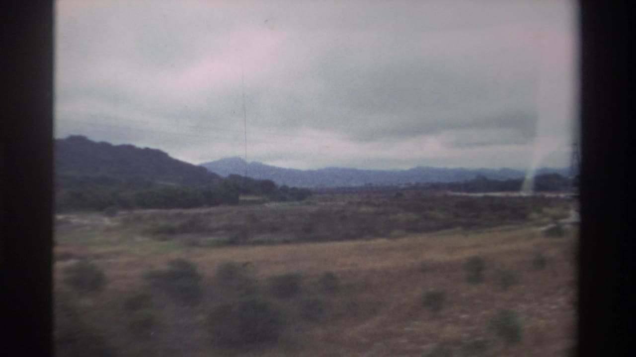 8mm home movie, 1982 Taiwan — open dry fields and distant mountains from a moving vehicle window, with visible film grain and faded color.