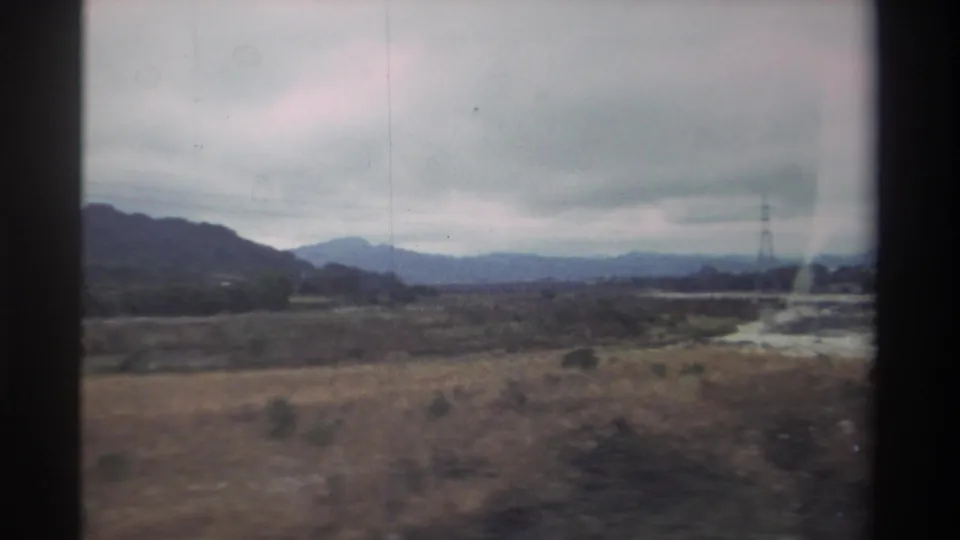 8mm home movie, 1982 Taiwan — a power transmission tower above dry scrubland with a mountain ridge behind, shot from a moving vehicle.