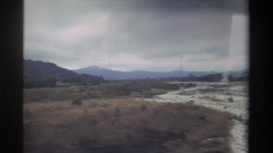 8mm home movie, 1982 Taiwan — sandy ground or dry channel in the foreground, transmission tower in midground, mountains behind, shot from a moving vehicle.