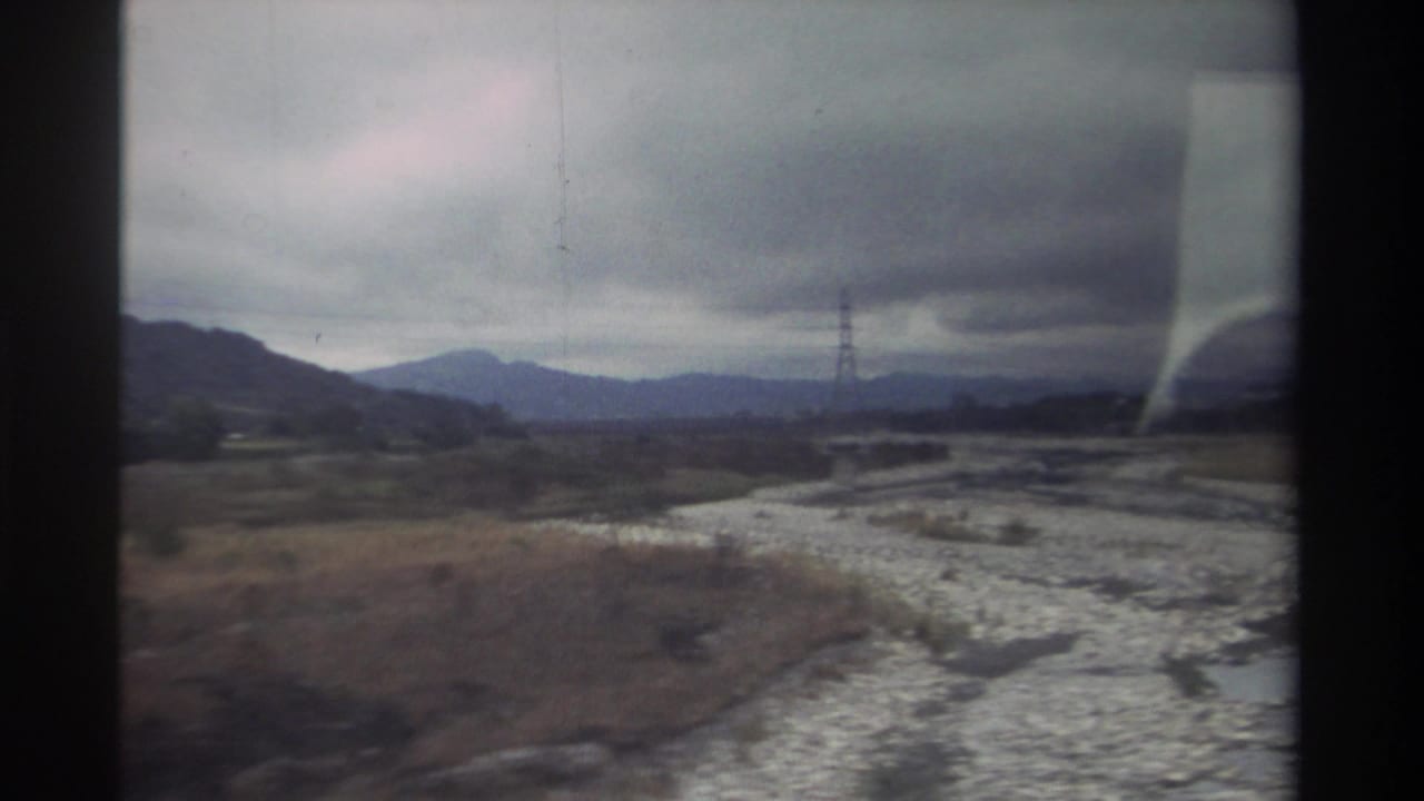 8mm home movie frame from Taiwan, circa 1982 — wide rocky riverbed, distant mountain range, lattice transmission pylon, and overcast sky shot from a vehicle window.