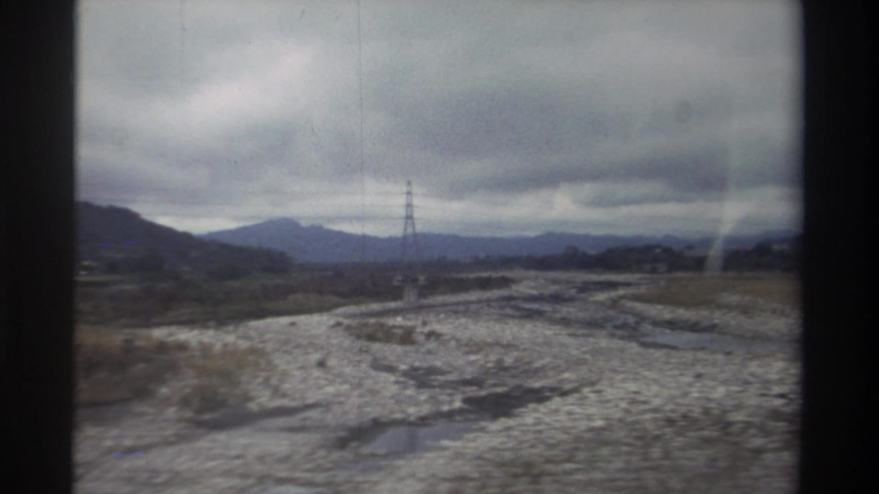 8mm home movie from Taiwan, circa 1982 — vehicle-window view of braided rocky riverbed, lattice pylon now at right, and distant mountains under an overcast sky.