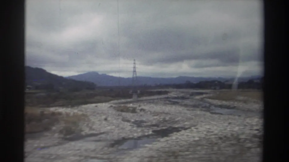 8mm home movie from Taiwan, circa 1982 — vehicle-window view of braided rocky riverbed, lattice pylon now at right, and distant mountains under an overcast sky.