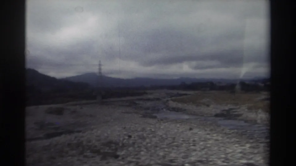 8mm home movie from Taiwan, circa 1982 — expansive overcast sky over rocky riverbed with lattice pylon at left and distant mountain silhouette, vehicle perspective.
