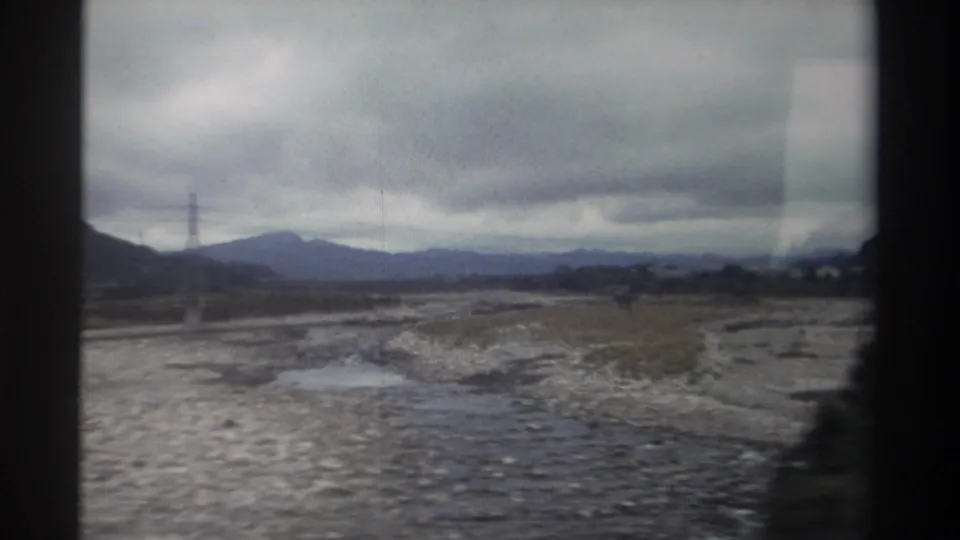 8mm home movie from Taiwan, circa 1982 — rocky braided riverbed with water reflections, lattice pylon, distant mountain range, and dark foliage at frame edge.