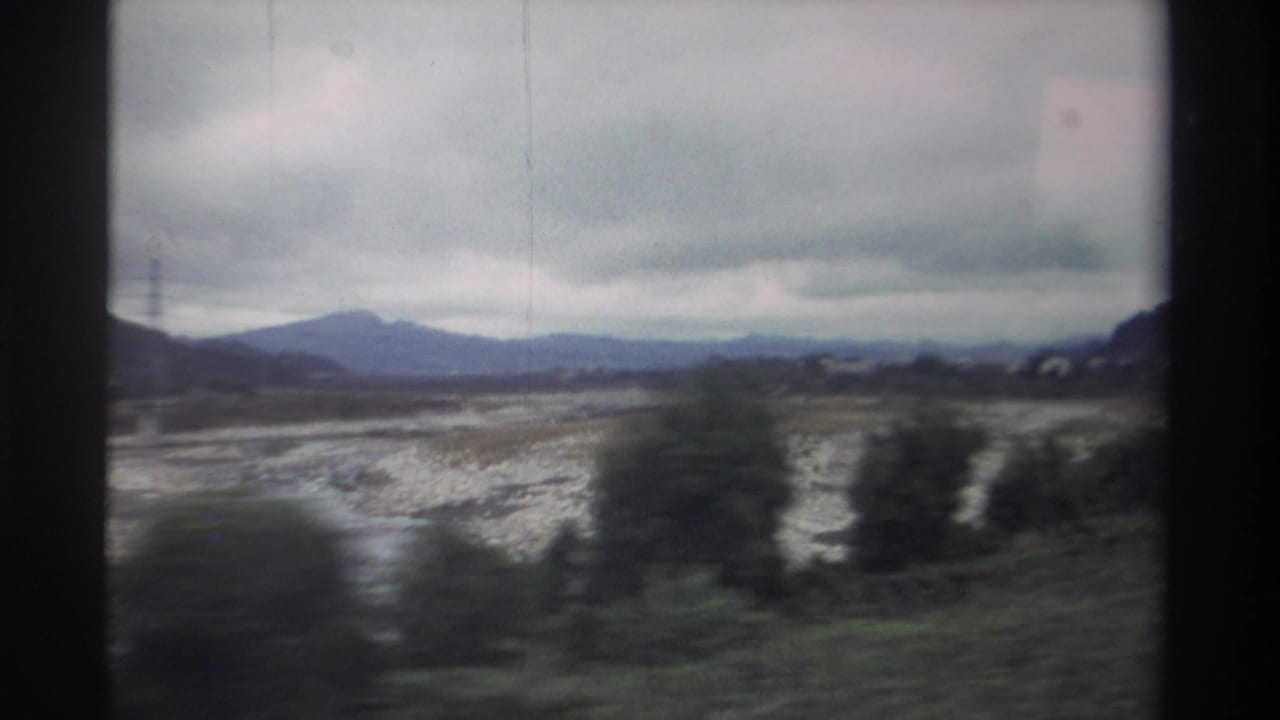 Overcast rural landscape from a moving vehicle: wide dry riverbed with pale gravel, scattered trees, and distant mountains.