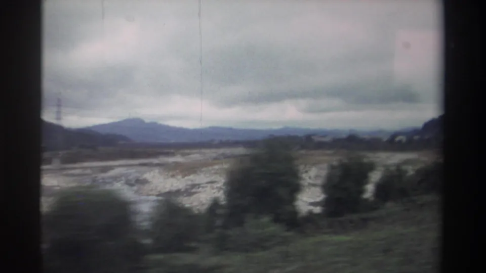 1982 Taiwan countryside shot from a moving vehicle — wide pale dry riverbed, mid-ground trees, and a mountain range under an overcast sky on 8mm film.