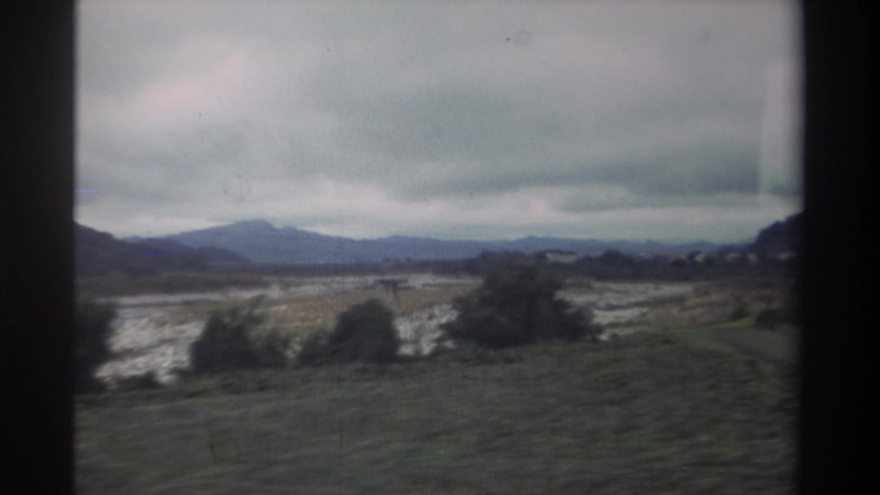 Pale dry riverbed and mid-ground treeline with mountain range in background under heavy cloud cover, shot from a moving vehicle.
