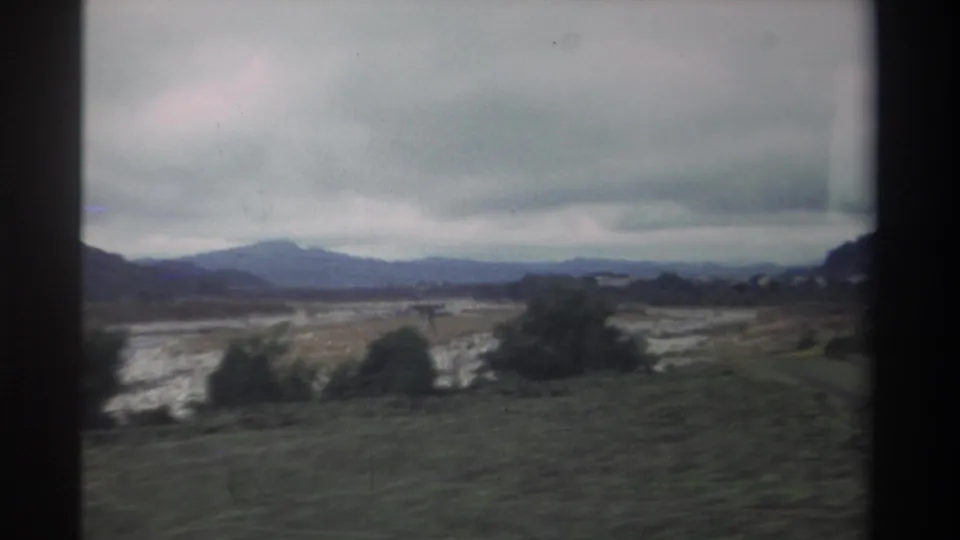 Continuing vehicle-shot view of a wide pale dry riverbed, a mid-ground tree line, and distant mountains in Taiwan's countryside, 1982, on 8mm film.