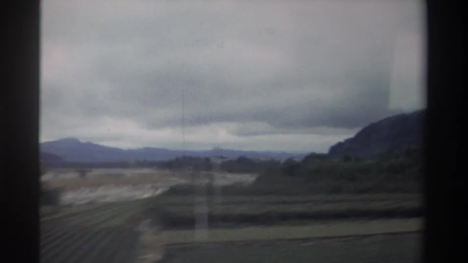 Flat agricultural plain and low rocky hills in Taiwan, 1982, with a prominent vertical film-scratch artifact crossing the frame, shot on 8mm.