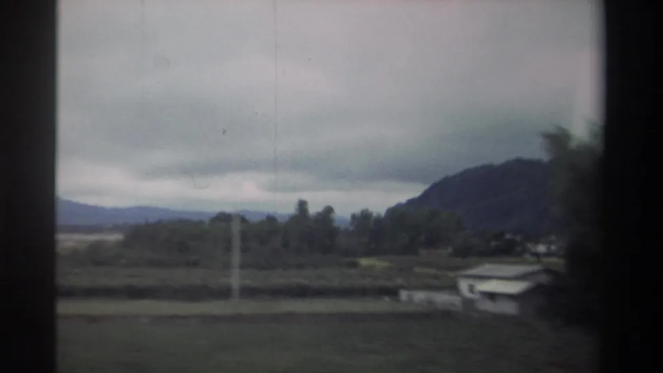 Rural Taiwan countryside in 1982 — flat fields, a rocky hill, small low-rise structures, a utility pole, and trees on the right, captured on 8mm from a moving vehicle.