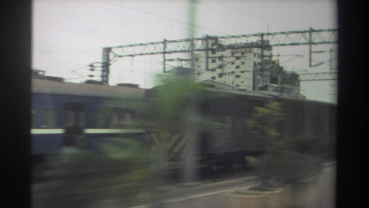 8mm frame from a moving train in Taiwan, 1982: freight wagons blur past, a blue passenger car edges into frame, and catenary gantries cross an overcast sky.