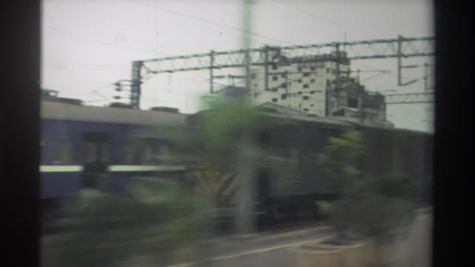 8mm frame from a moving train in Taiwan, 1982: freight wagons blur past, a blue passenger car edges into frame, and catenary gantries cross an overcast sky.