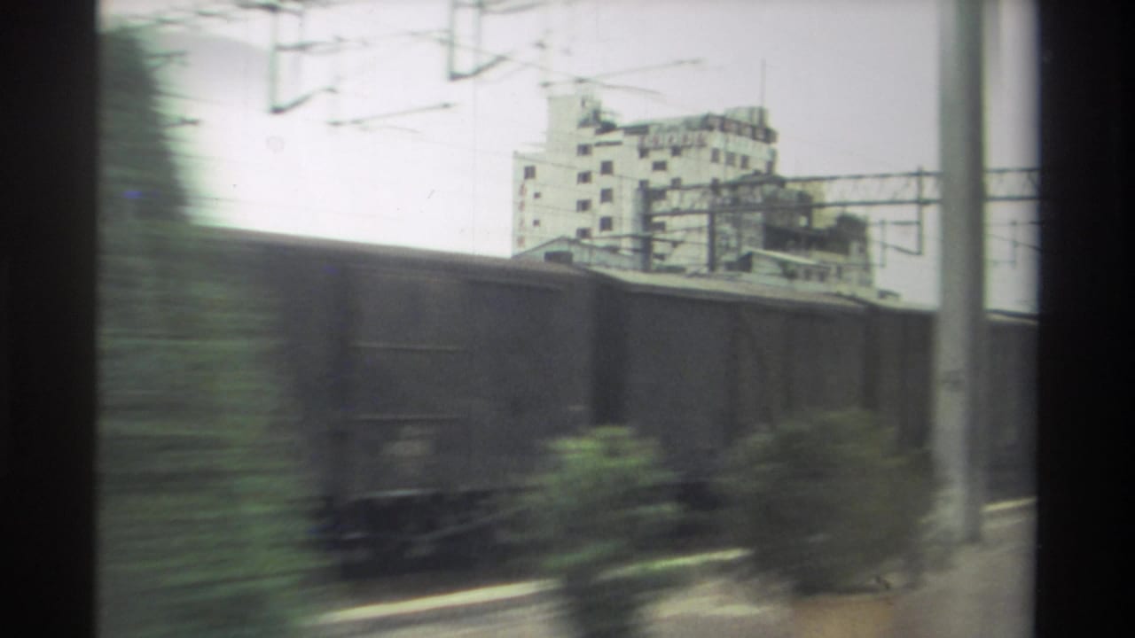 8mm home movie frame, Taiwan 1982: freight wagons dominate the foreground with lateral motion blur; catenary gantries and a concrete building rise behind.
