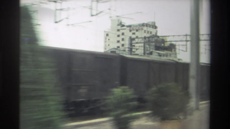 8mm home movie frame, Taiwan 1982: freight wagons dominate the foreground with lateral motion blur; catenary gantries and a concrete building rise behind.