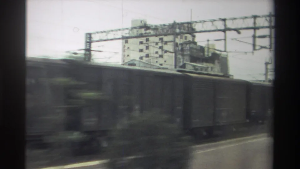 1982 Taiwan 8mm footage: the vegetation clears to reveal catenary gantry infrastructure, dark freight wagons, and a concrete urban building across the rail yard.