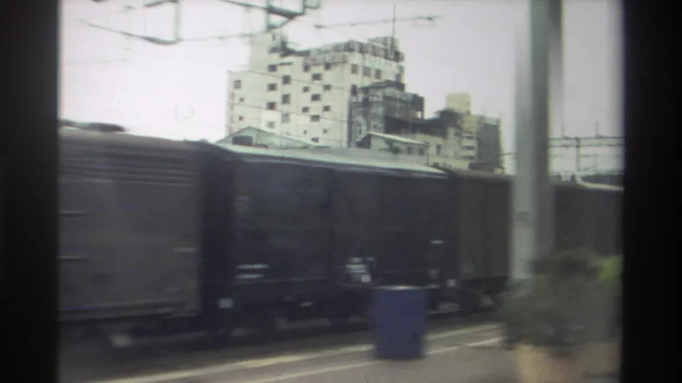 Final 8mm frame, Taiwan rail yard 1982: freight wagons dominate with a blue drum on the platform edge, catenary wires above, and a concrete building at upper right.