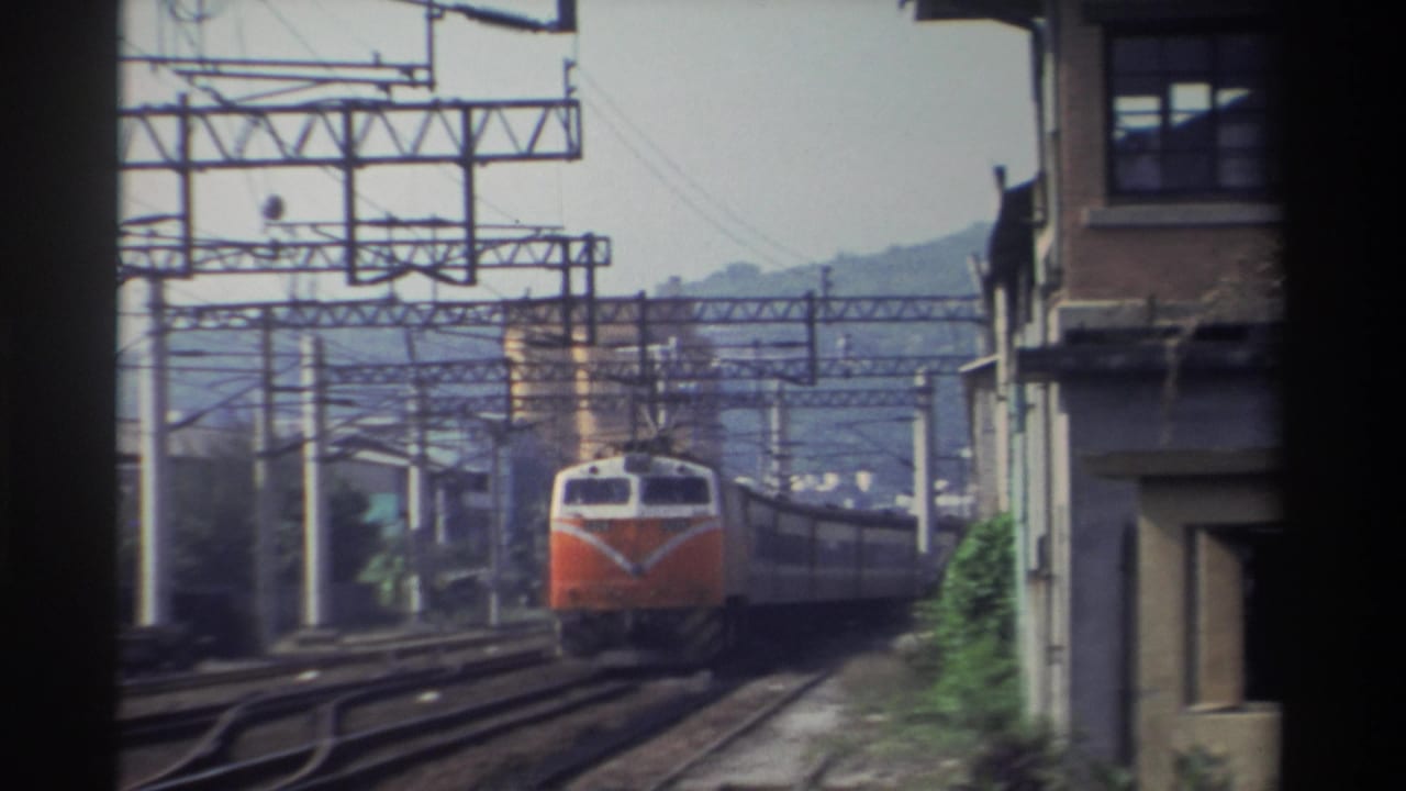 Orange-and-white Taiwan Railways locomotive approaching head-on, electric catenary gantry structures prominent, multi-story brick building at right, hazy mountains behind, Taiwan, 1982.