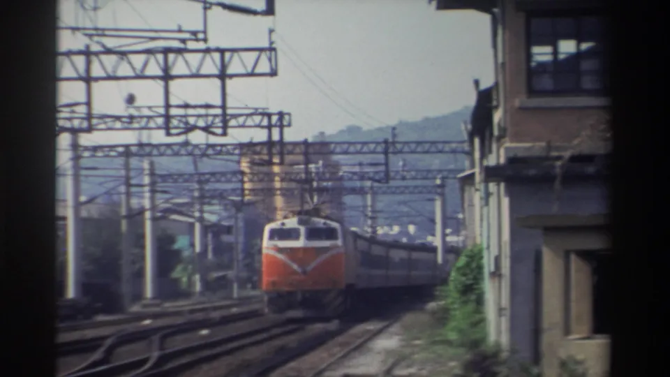 Orange-and-white Taiwan Railways locomotive approaching head-on, electric catenary gantry structures prominent, multi-story brick building at right, hazy mountains behind, Taiwan, 1982.