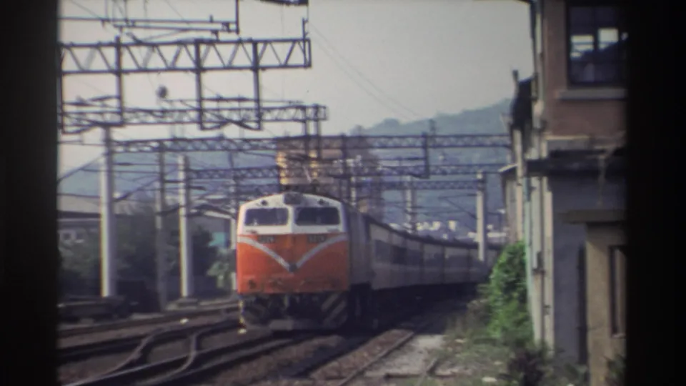 Orange-and-white Taiwan Railways locomotive approaching closely with white chevron emblem and number plate visible on nose, catenary poles and weathered building at right, mountains behind, Taiwan, 1982.