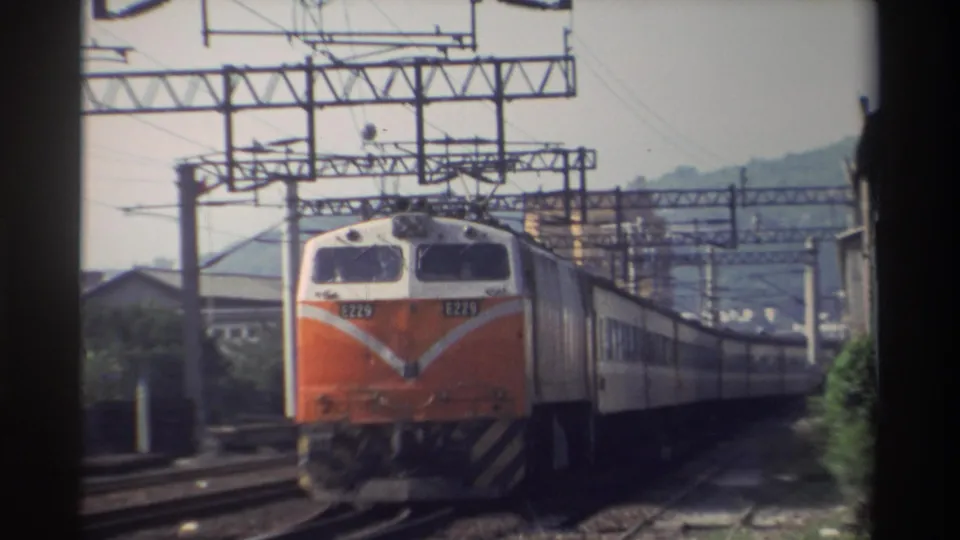 Close frontal view of orange-and-white Taiwan Railways locomotive with legible number plate and white chevron nose emblem, catenary wires overhead, weathered building at far right, Taiwan, 1982.