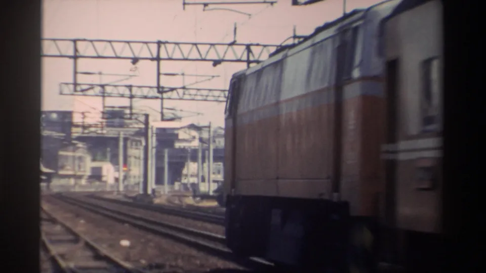 Taiwan Railway E229 electric locomotive in orange livery seen from trackside, catenary gantries and low urban buildings in background, circa 1982.