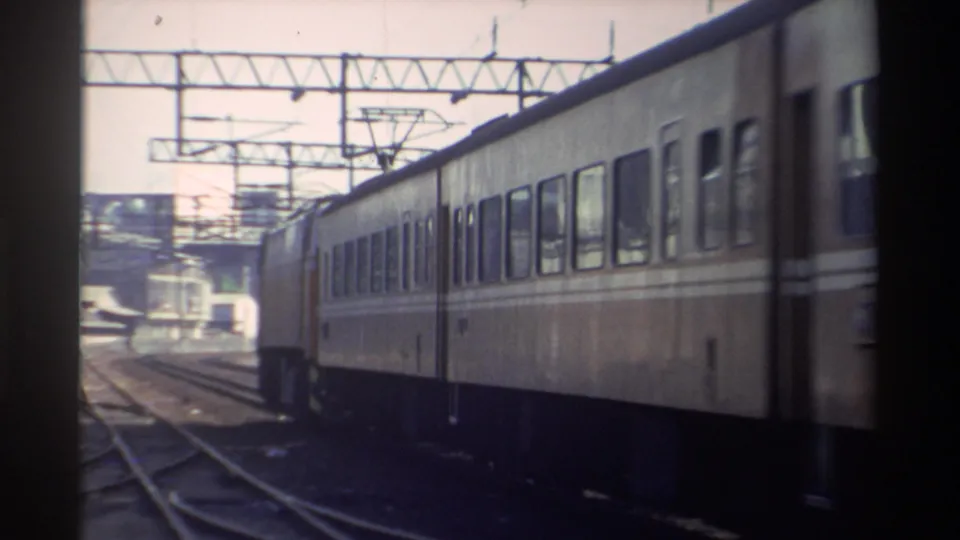 A cream-and-brown electric rail car passes through a Taiwan track area in bright daylight, with catenary structures overhead.