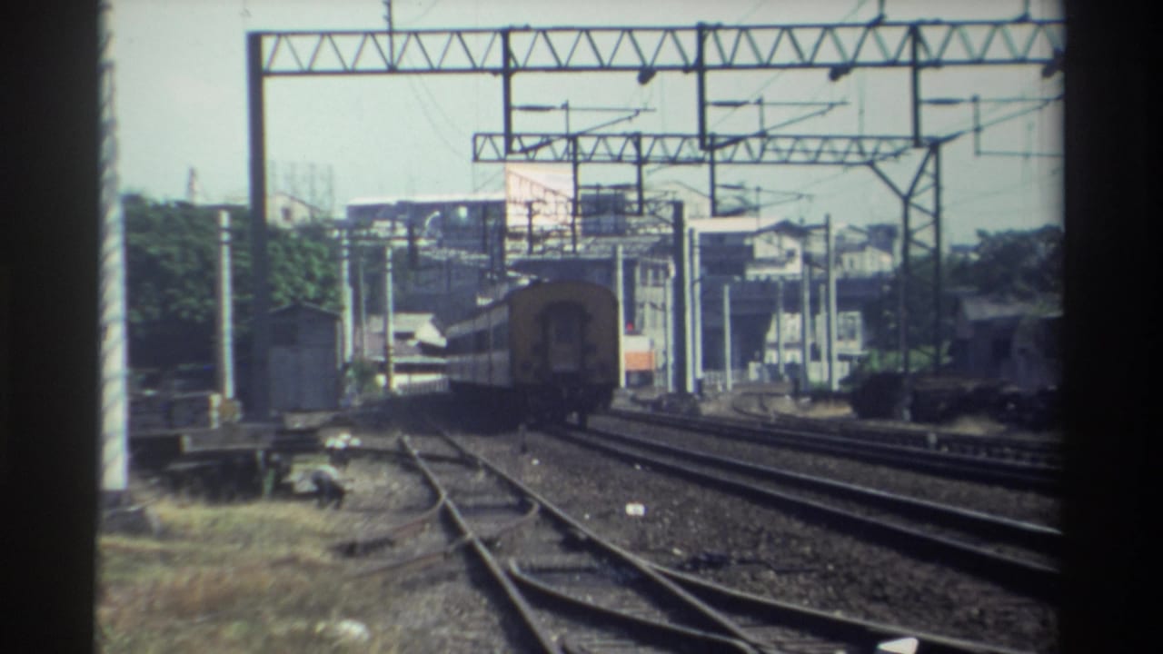 Train receding on rail lines with overhead gantries and buildings in the background.