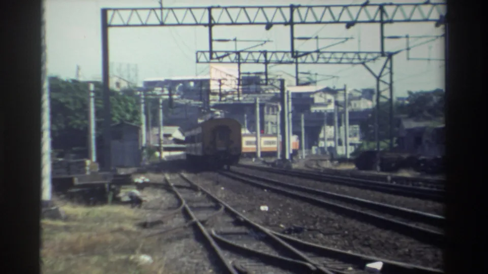 A distant train moves along the tracks while another cream-and-orange railcar appears behind the catenary structures in Taiwan.