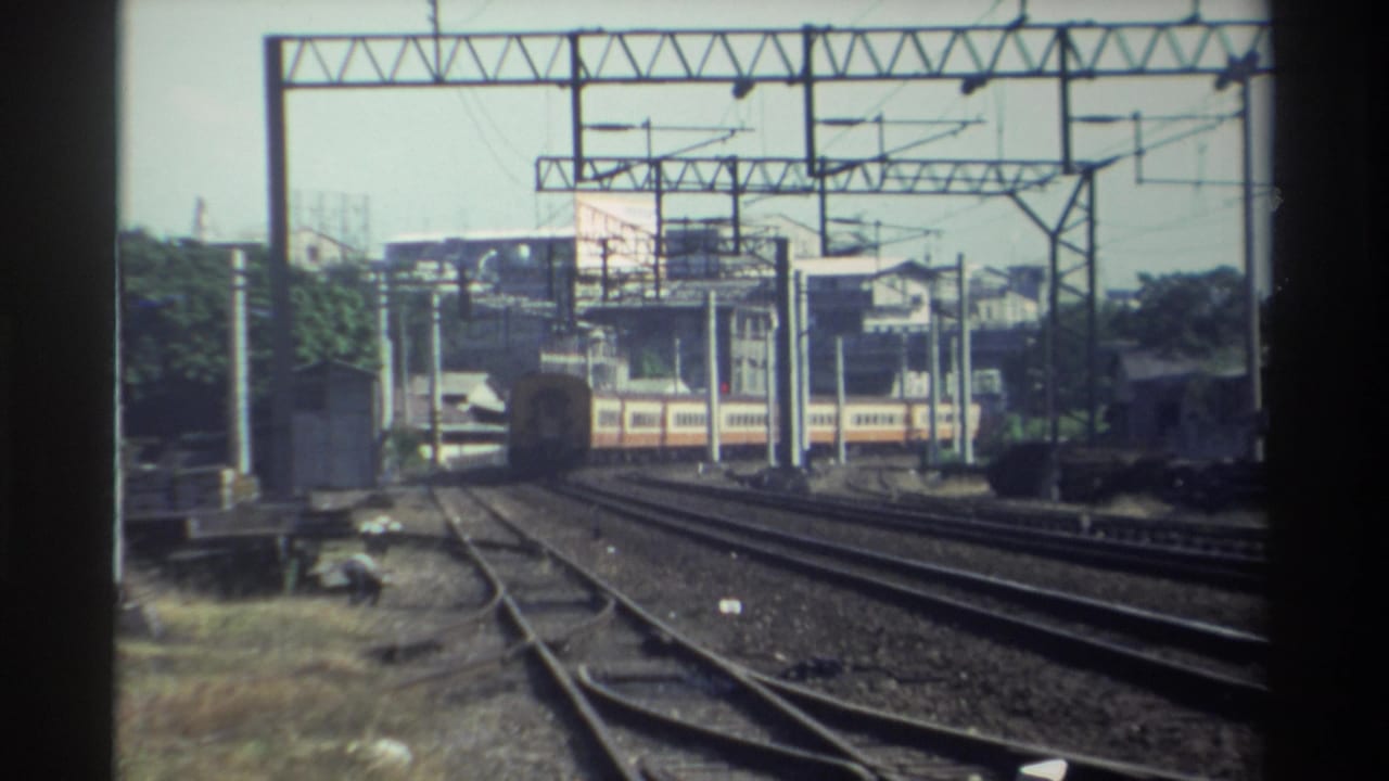 The passenger train continues through the Taiwan rail yard, framed by gantries, poles, ballast, trees, and distant buildings.
