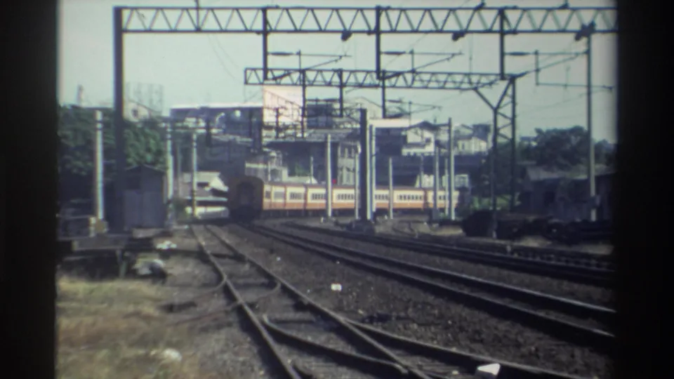 A vintage 8mm view of orange-and-cream train cars moving across parallel tracks in Taiwan, with overhead rail equipment above.