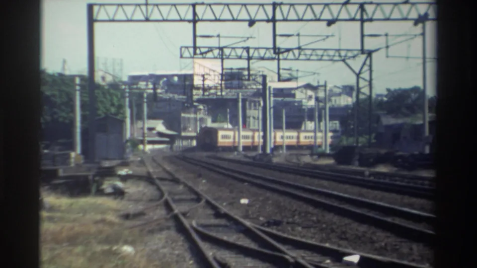 1982 Taiwan rail scene with a departing passenger train, multiple tracks, overhead power structures, and low-rise development beyond.